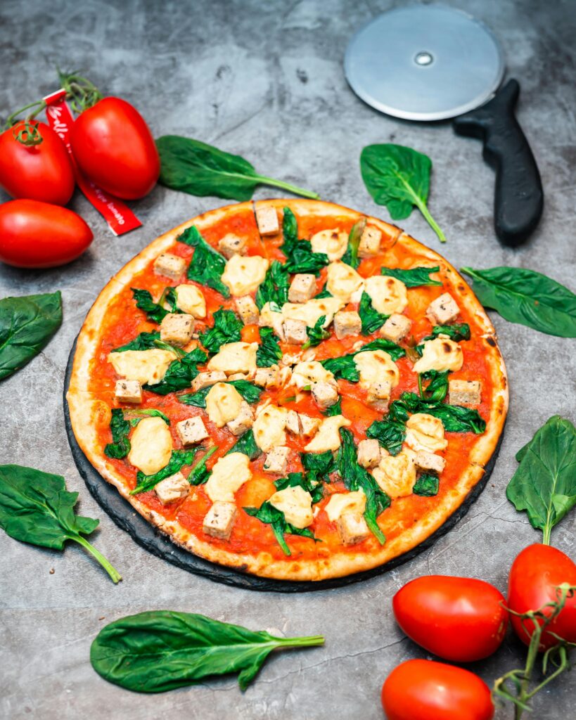 Bright and fresh pizza topped with tofu, spinach, and mozzarella surrounded by tomatoes and spinach leaves.